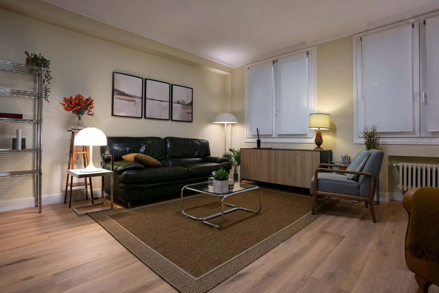 Sala de estar moderna con sofá de cuero negro, mesa de centro de vidrio, lámpara de pie y estantería metálica. Decoración minimalista con cuadros en la pared y plantas. Suelo de madera clara y alfombra de yute.